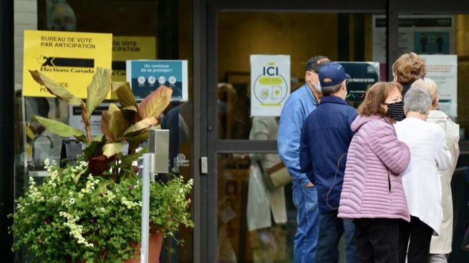 Des personnes font la queue devant l'entrée d'un bâtiment sur laquelle est collée une affiche jaune indiquant Bureau de vote par anticipation.