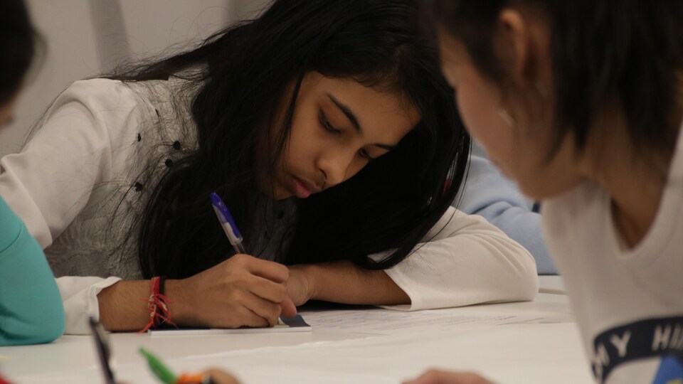 Une fille prend des notes sur un calepin à une table entourée d'autres jeunes. 