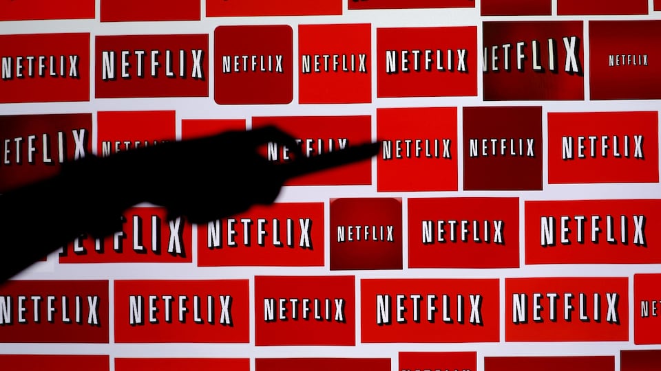 L'ombre d'une main tenant une télécommande, devant de nombreux logos de Netflix.