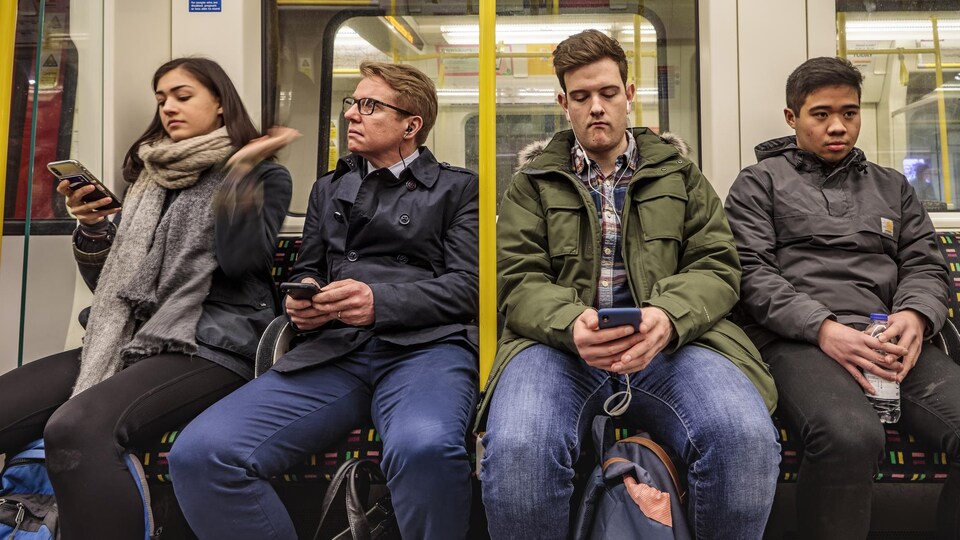 Quatre passagers assis côte à côte sur une banquette de métro. Trois d'entre eux tiennent leur téléphone entre leurs mains.