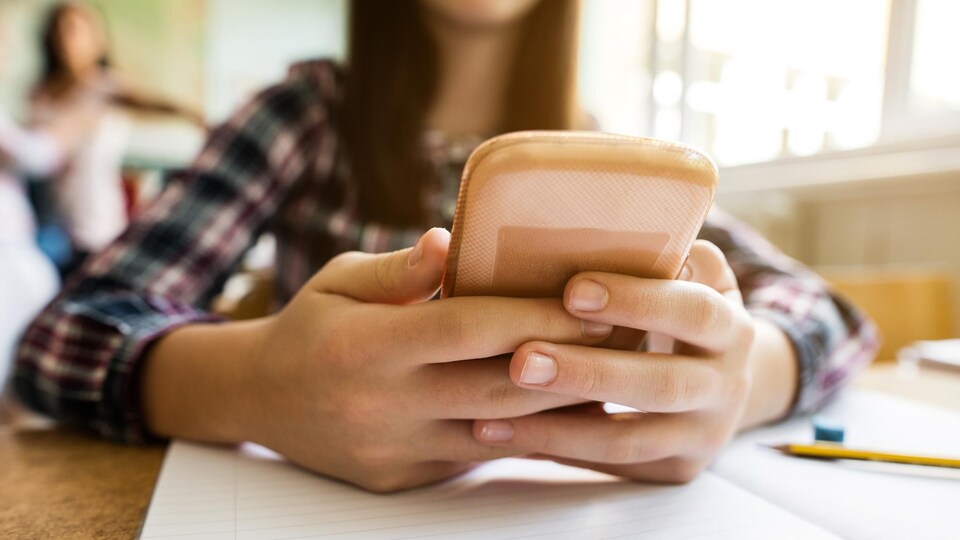 Un gros plan sur les mains d'une jeune fille qui tient un téléphone intelligent. Ses mains sont posées sur son pupitre d'école, par dessus un cahier ligné.