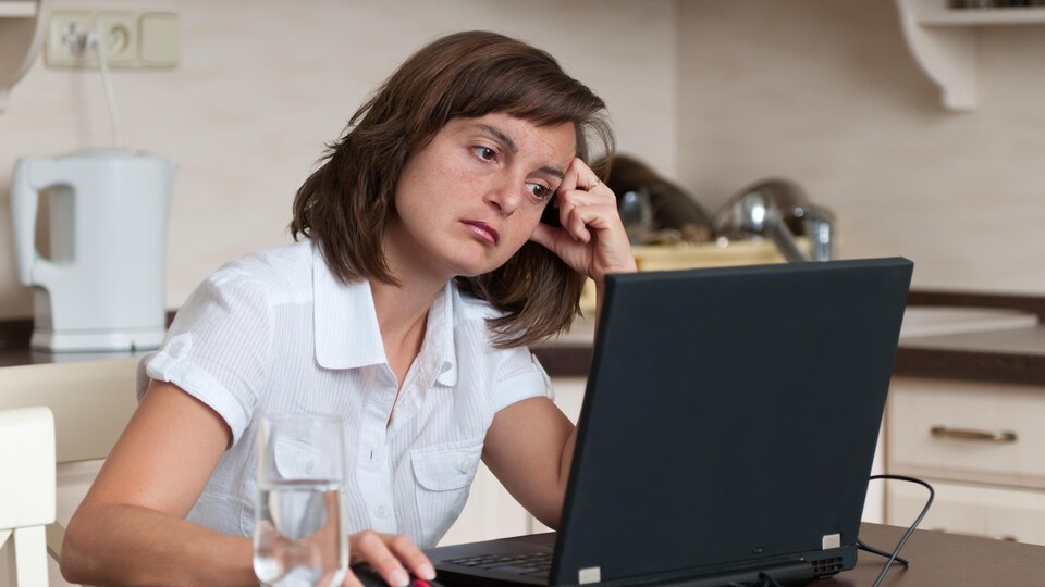 Une femme assise dans sa cuisine semble s'ennuyer devant son écran d'ordinateur.