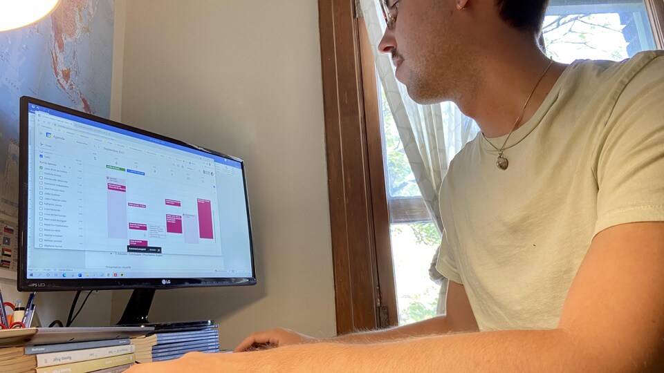 Un homme est assis chez lui devant son écran d'ordinateur. Il regarde son horaire pour la semaine à venir. 