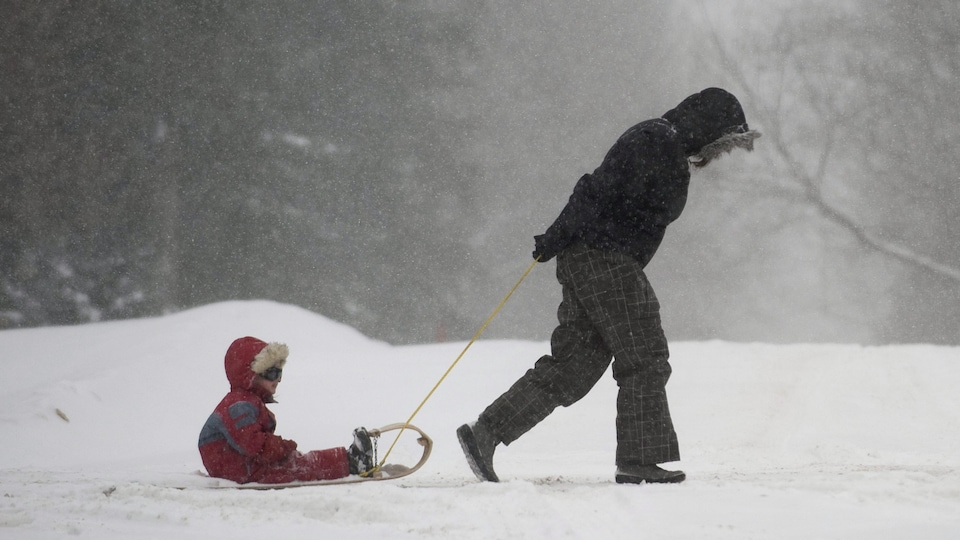 Une femme traîne un enfant, assis sur un traîneau, en pleine tempête de neige.