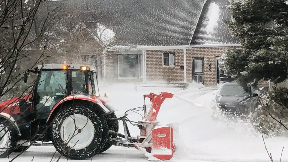 Un tracteur de déneigement souffle la neige devant une maison.