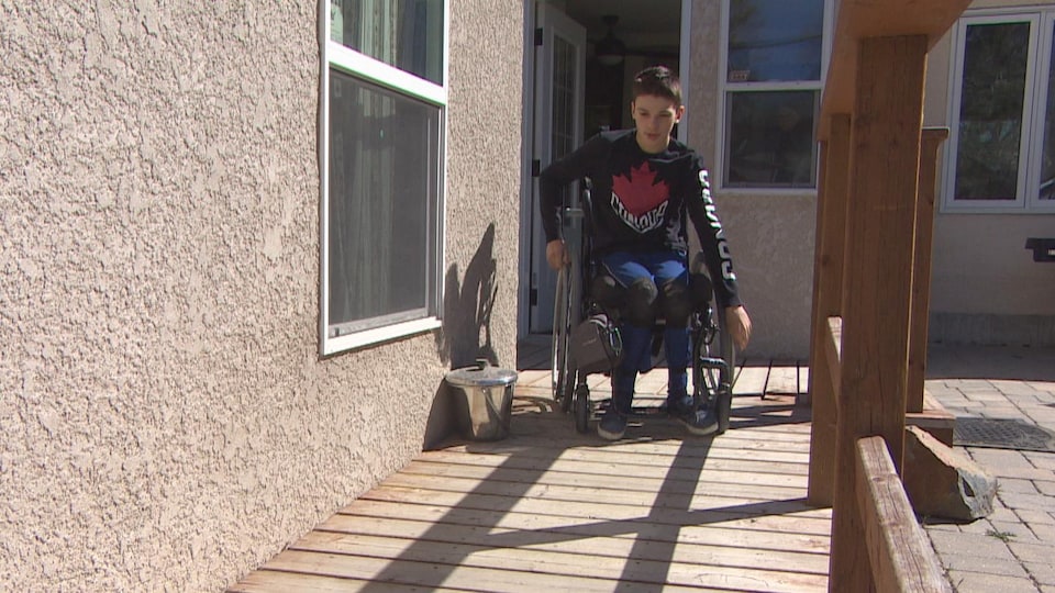 Un jeune homme dans un fauteuil roulant, sur une rampe d'accès à l'extérieur d'une maison.