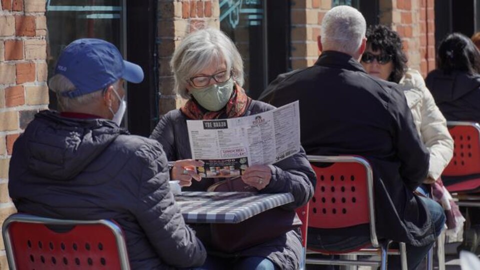Un dame attablée sur une terrasse avec un homme consulte un menu. Tous deux portent le masque.
