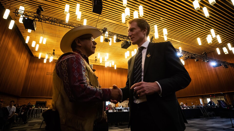 Sous un éclairage tamisé dans une grande salle de conférence, le ministre en complet serre la main du chef qui porte un chapeau de cowboy.