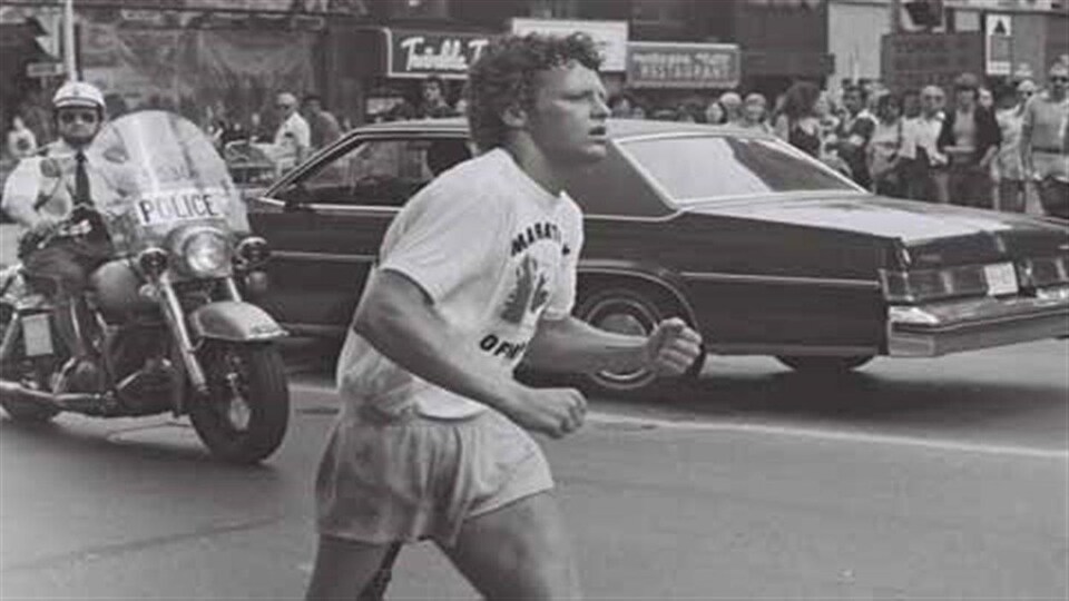 Un coureur avec une jambe artificielle court dans une rue alors qu'il est escorté des policiers. La photo est en blanc et noir et date des années 1980.