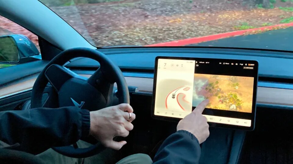 Un homme assis dans le siège conducteur tient le volant d'une Tesla de la main gauche et joue à un jeu sur un écran tactile de la main droite.