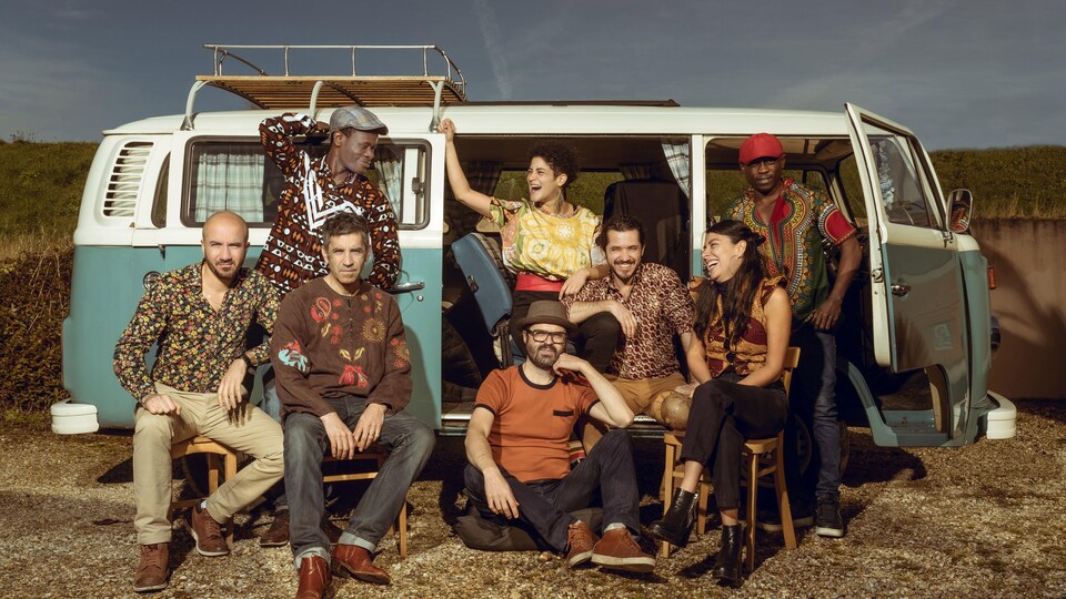 Les musiciens et musiciennes s'esclaffent devant une petite fourgonette aux portes ouvertes. 