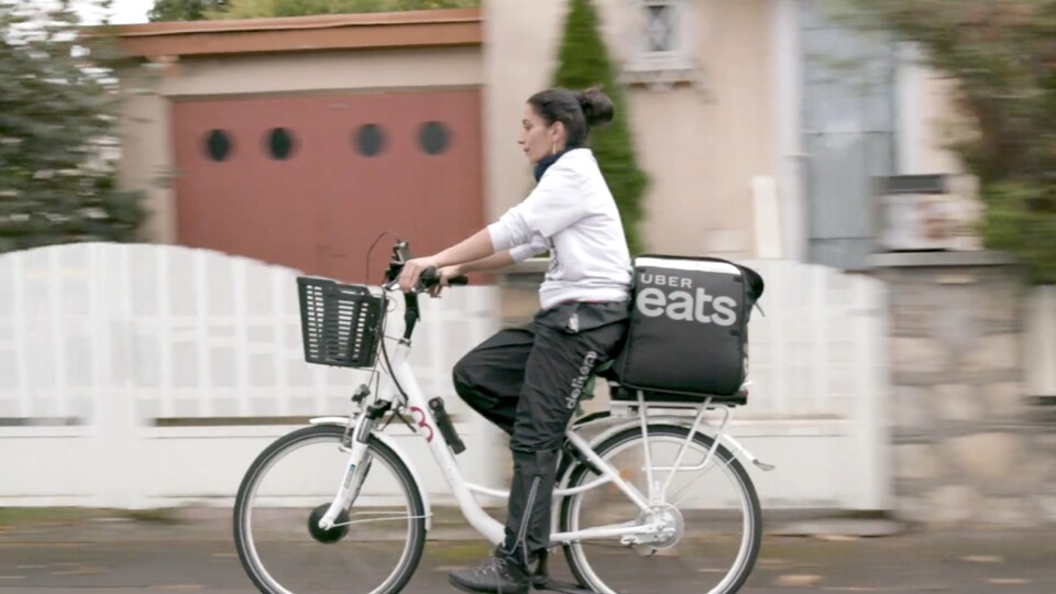 une femme sur un vélo, un sac de livraison UBER EATS à l'arrière, un pantalon DELIVEROO