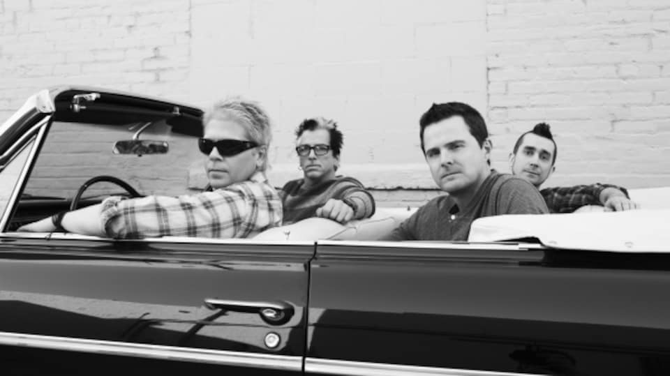 Photo en noir et blanc de quatre hommes dans une voiture décapotable qui regardent vers l'appareil photo.