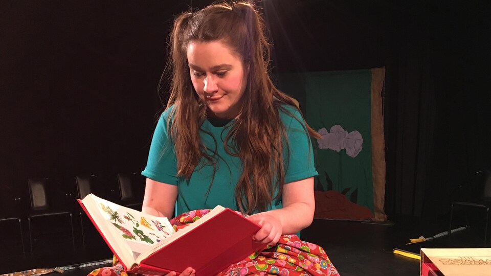 Une jeune fille avec des couettes assise sur une table avec un livre ouvert posé sur les genoux. 