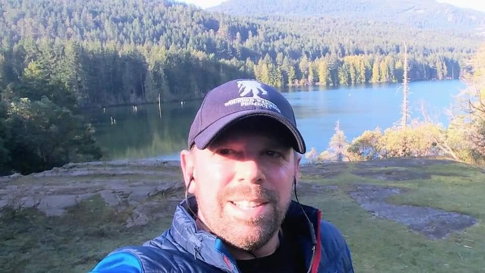 Un homme porte une casquette et un manteau sport bleu et a de petits écouteurs dans les oreilles. Il est à l'extérieur devant un lac entouré d'une forêt immense.