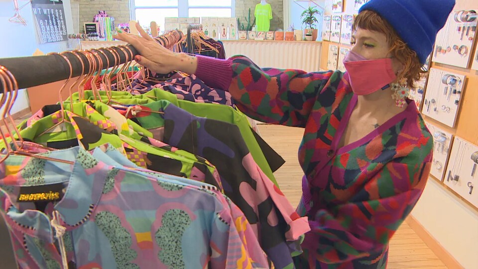 Une femme regarde des vêtements à l'intérieur d'une boutique et porte un masque sur le visage.