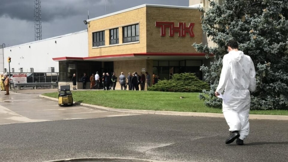 Un homme en combinaison blanche et des gens rassemblés à l'entrée d'une usine.