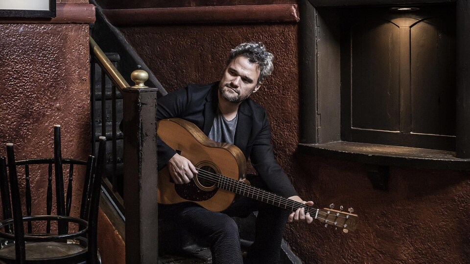 Portrait en couleur de Thomas Hellman, assis sur les marches d'un escalier, guitare à la main