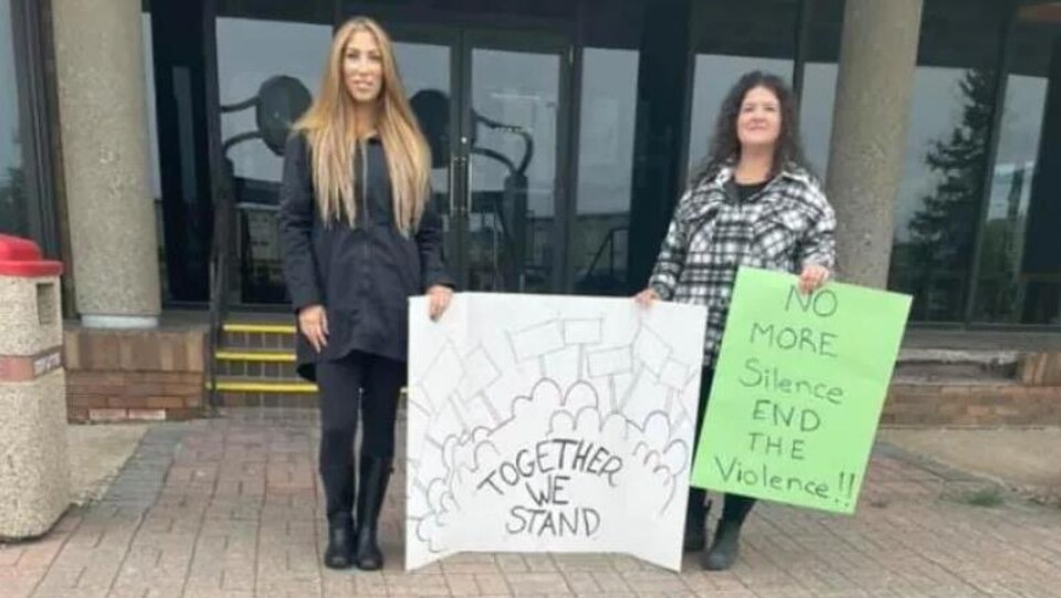 Deux femmes, debout, tiennent une pancarte avec des inscriptions.