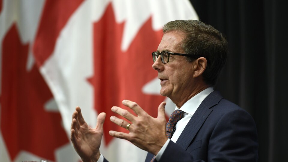Tiff Macklem parle, avec un drapeau du Canada en arrière-plan.