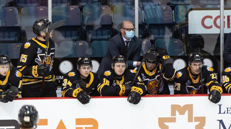 Un entraîneur de hockey avec un masque derrière ses joueurs au banc.