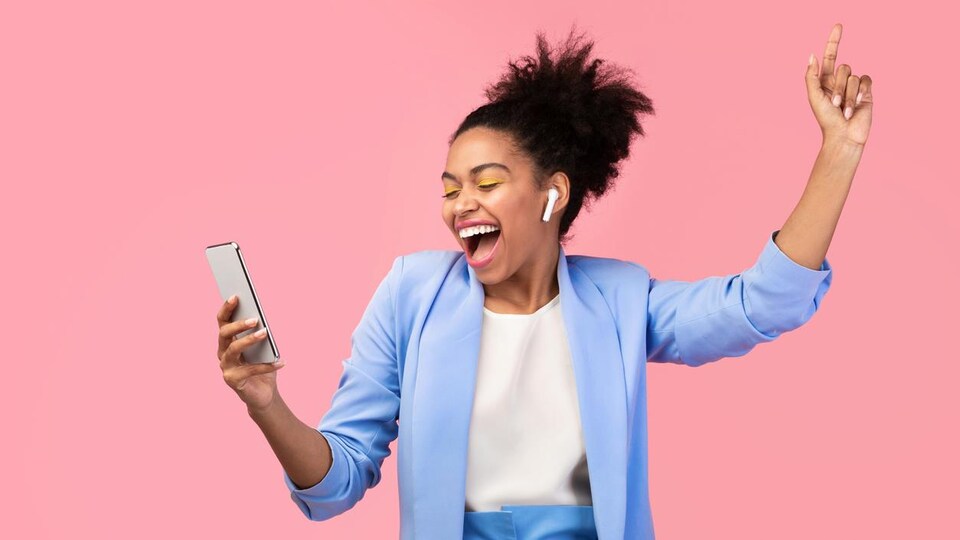 Une femme souriante écoute de la musique avec des écouteurs sans fil sur son téléphone en dansant devant un fond rose.