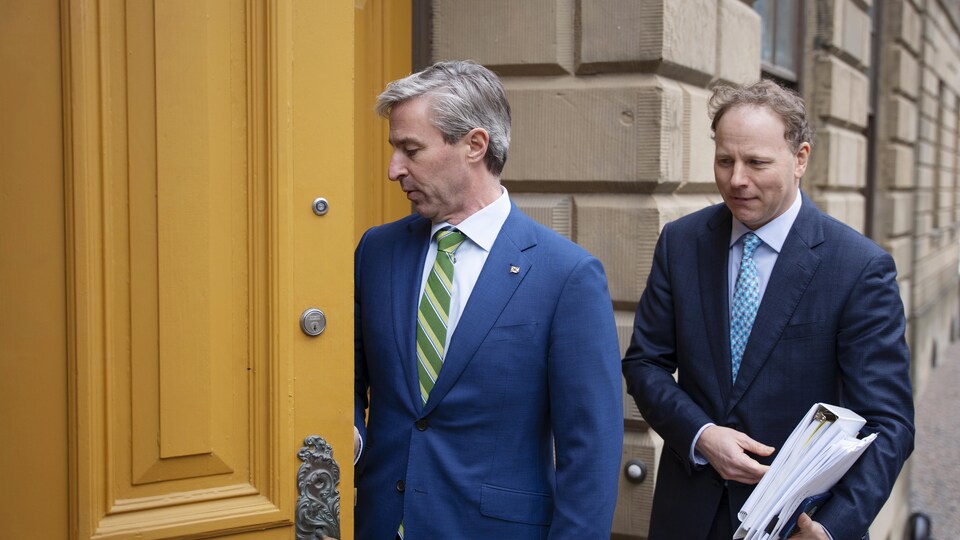 Tim Houston ouvre la grande porte en bois de Province House. Allan MacMaster marche derrière lui avec un cartable et une pile de documents dans les mains.