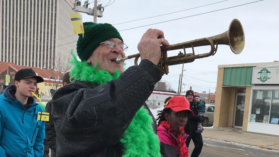 Une personne porte une écharpe à plumes vertes et souffle à la trompette.