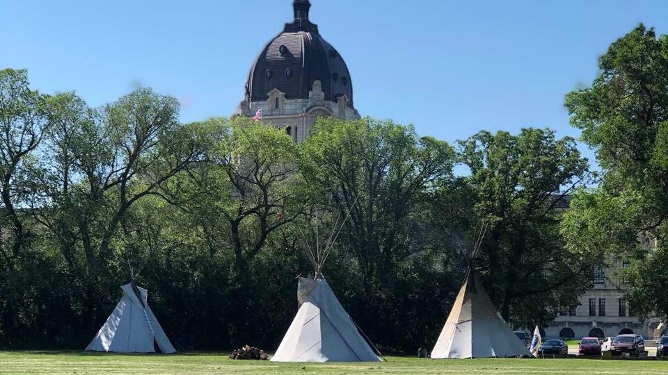 Trois tipis installés sur un gazon devant un boisé.Une partie de l'assemblée législative de la Saskatchewan est visible derrière le boisé.