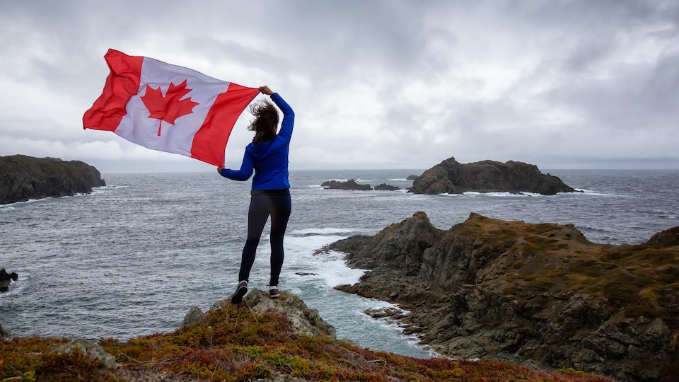 Une femme debout sur la berge agite un drapeau du Canada près de l'océan.