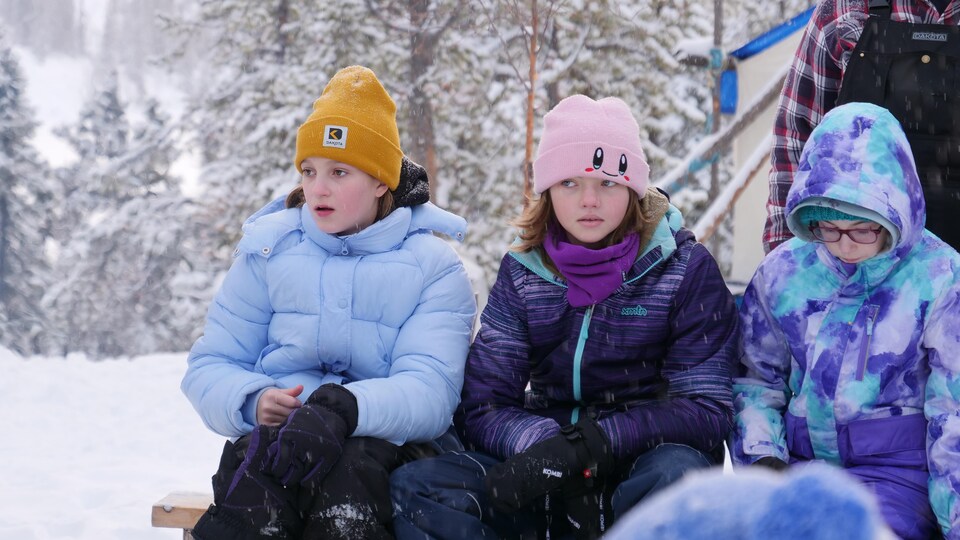 Trois enfants sont assis dehors, ils portent une tuque et un manteau d'hiver, devant des arbres recouverts de neige.