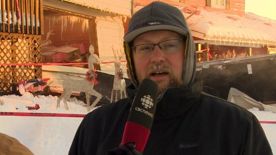 Personne avec tuque et manteau tenant un micro sur lequel est écrit CBC News, devant une maison et des décorations de Noël, le 6 décembre 2022, à Yellowknife.