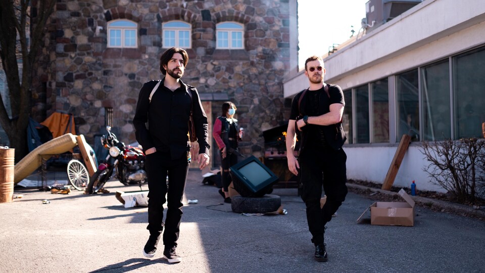 Yannick De Martino et Alexandre Bisaillon marchent près d'un bâtiment abandonné devant lequel se trouvent des débris et une moto.