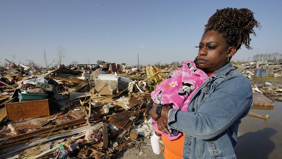 Une femme noire tient une fillette dans ses bras, devant les restes de maisons effondrées.
