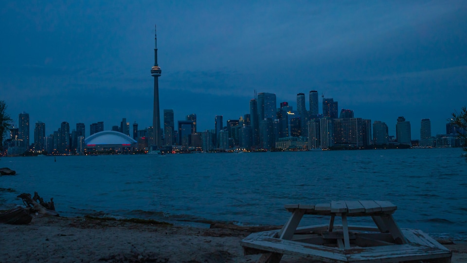 Le centre-ville de Toronto avec des dizaines de gratte-ciels illuminés à la tombée du jour vu d'une table de pique-nique d'une île sur le lac Ontario.