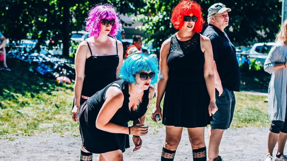 Trois femmes jouent en extérieur à la pétanque. Toutes les trois portent des perruques de couleur rouge, fuchsia et bleu, des lunettes de soleil et une robe noire.