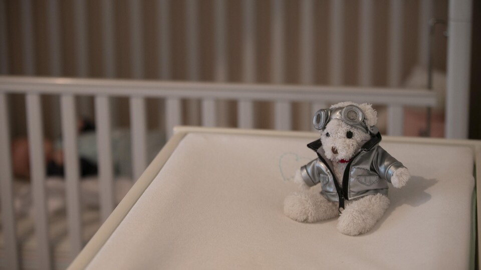 Un toutou vêtu d'un blouson aviateur et d'un casque sur une table avec en fond de décor un enfant dans son lit de bébé.