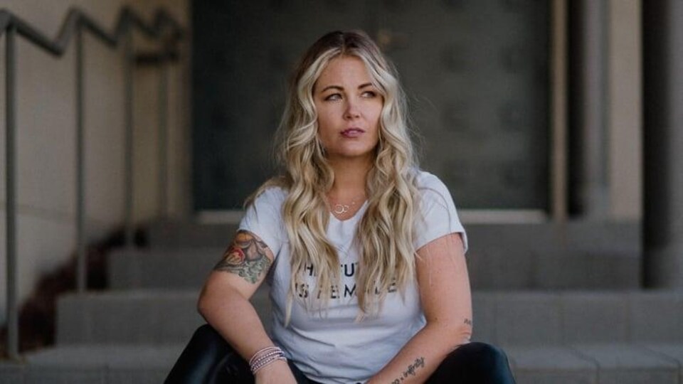 Une femme avec de longs cheveux blonds assise sur des marches regarde vers la droite.