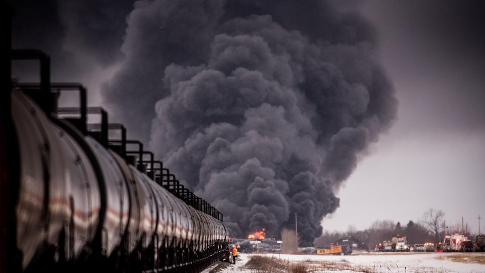 Plan longeant le train avec, au fond, les wagons en travers de la voie en feu avec un grand nuage de fumée qui monte dans le ciel.