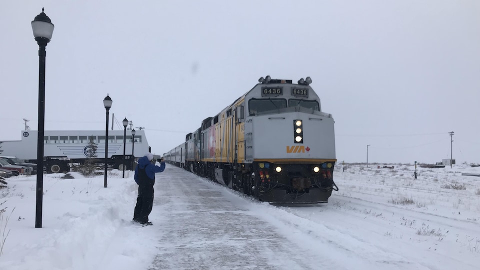 Une locomotive, vue de face, sur les rails enneigés. Sur le côté, un passant prend une photo du train.