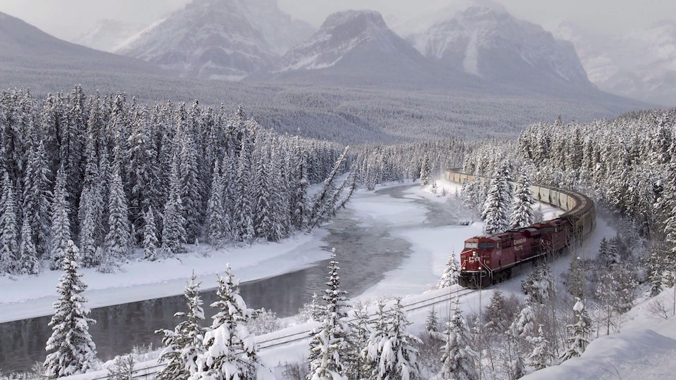 Un train rouge du  Canadien Pacifique roule au milieu de sapins recouverts de neige et le long d'une rivière à moitié gelée. Au loin, on voit les sommets enneigés des montagnes Rocheuses. 