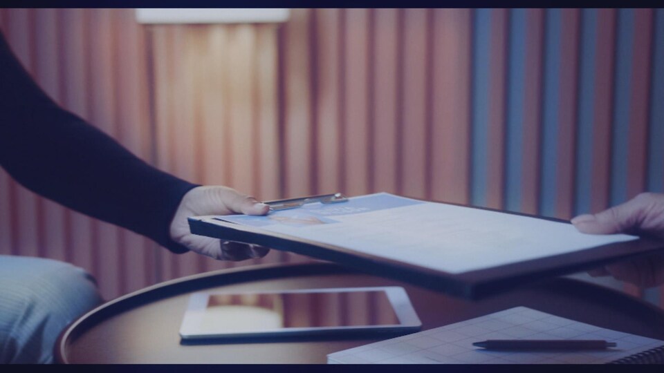 Deux personnes dont on ne voit que les mains s'échangent une tablette à laquelle est fixée un document papier.