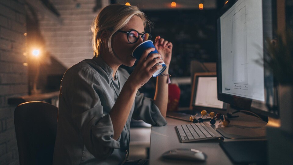 Une jeune femme boit un café en regardant un écran d'ordinateur assise à un bureau.
