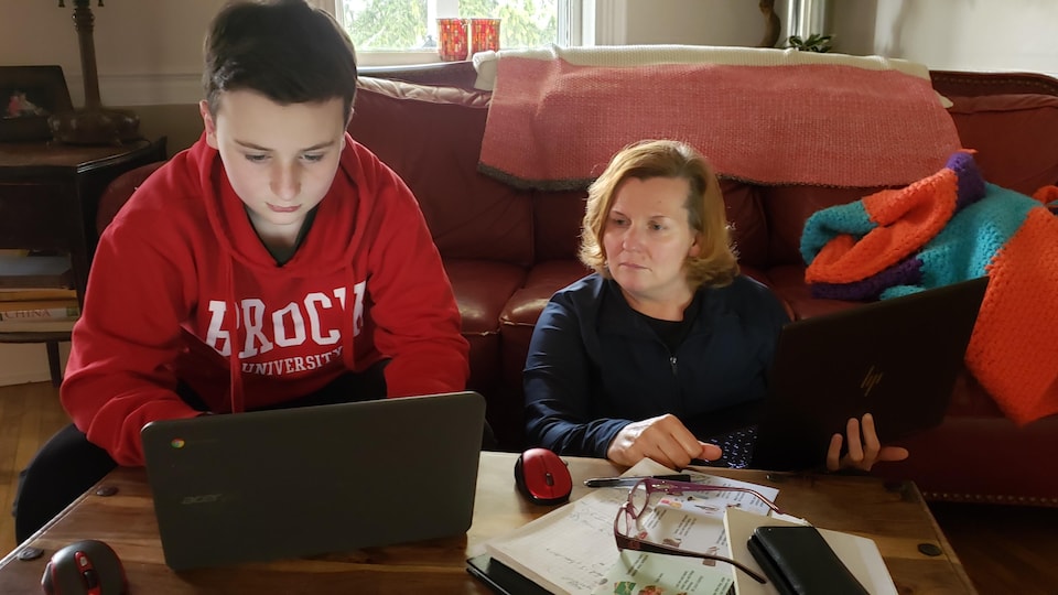 Une femme tient un ordinateur portable et regarde ce qu'un garçon fait sur un autre ordinateur. Ils sont assis sur le plancher d'un salon familial. 