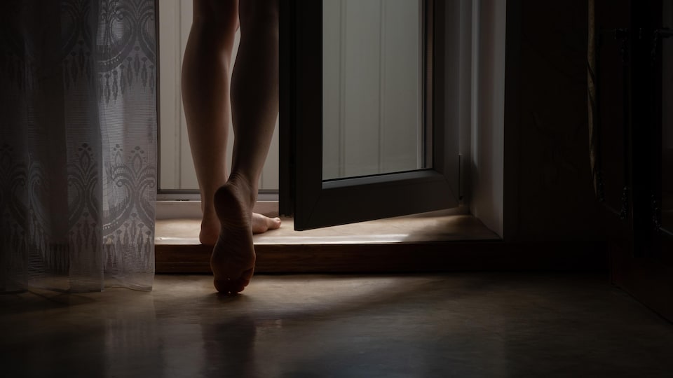 Une femme aux jambes nues, de dos, dans un appartement devant une porte entrouverte.