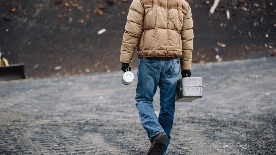 Un homme marche vers un chantier avec une boîte à lunch en métal à la main.