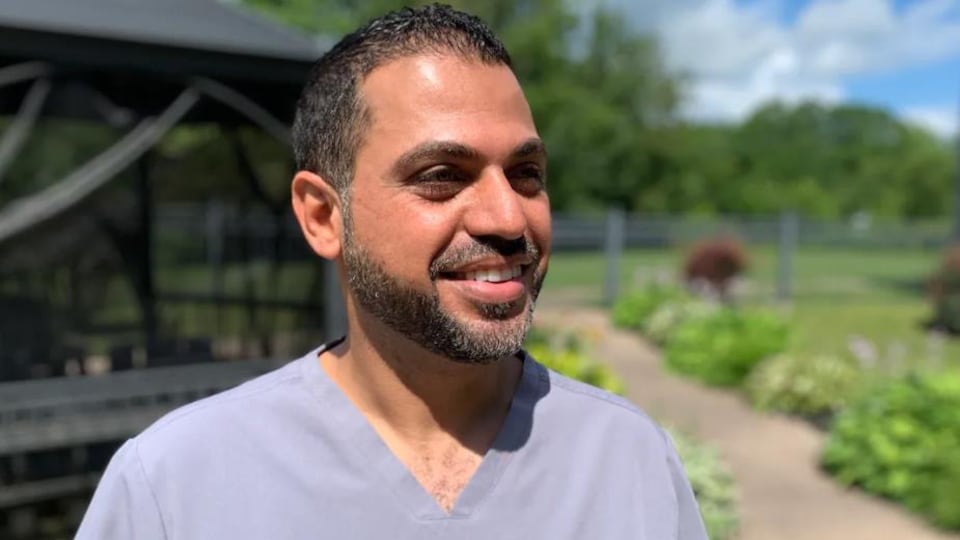 Un homme souriant en uniforme dans un jardin extérieur, sous le soleil.