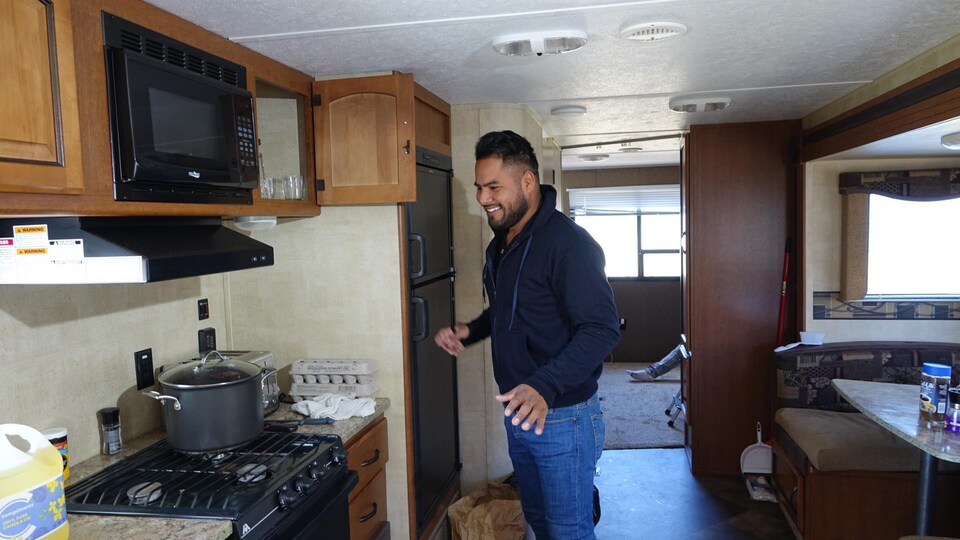 A smiling man stands in the kitchen of the trailer in which he lives.