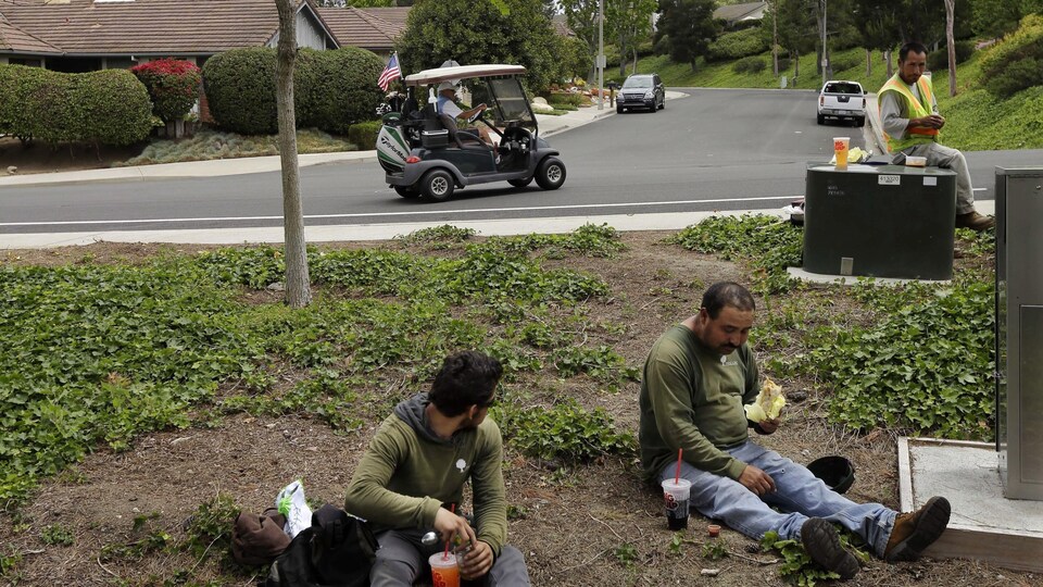 Des jardiniers se reposent assis par terre en regardant passer un homme fortuné dans une voiturette de golf.