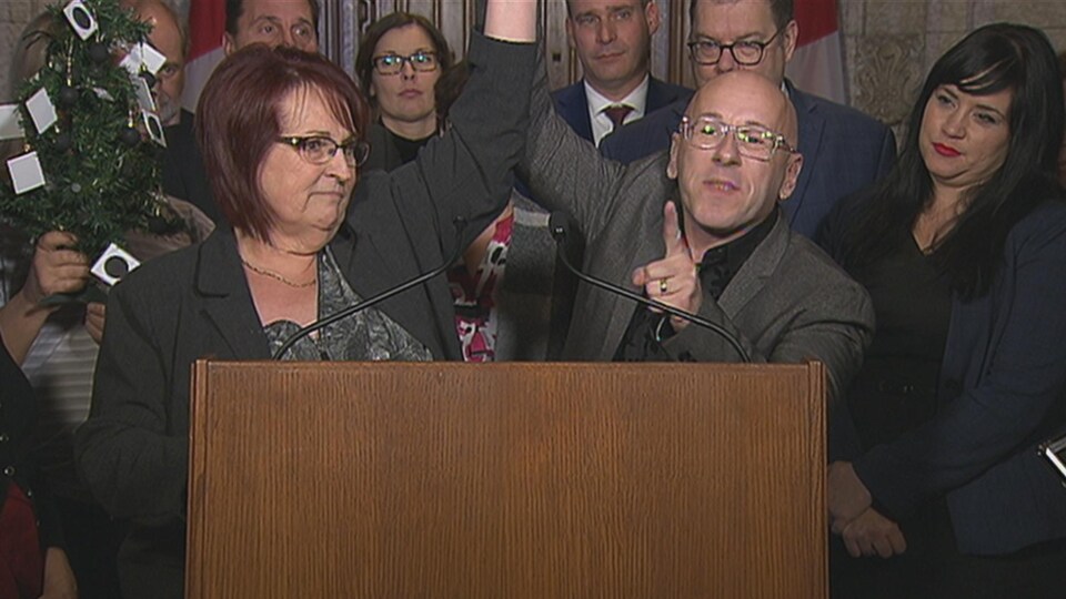 Un homme soulève le bras d'une femme, devant un podium, au Parlement du Canada.